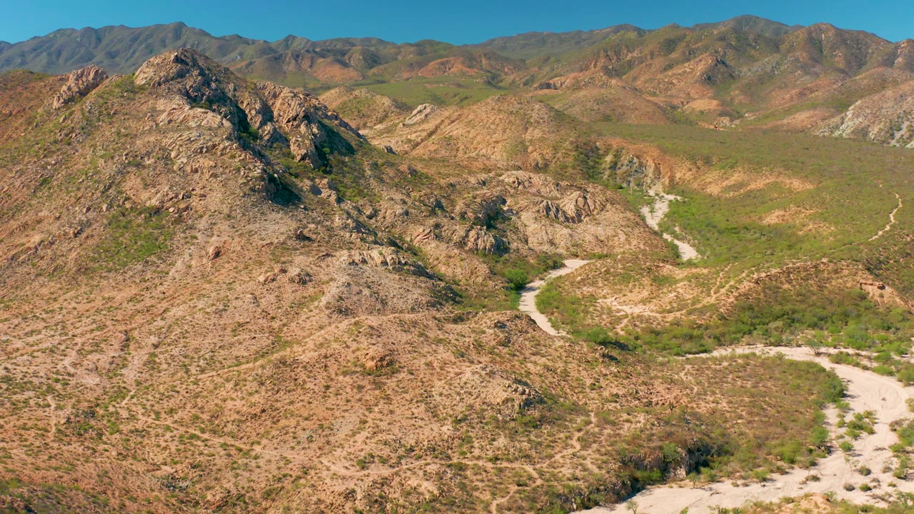 Cabo Pulmo aerial view with the dirt road forming a light curve near the lower right, tan slopes in the foreground, green vegetation in the valley, layered ridges, and no visible people.