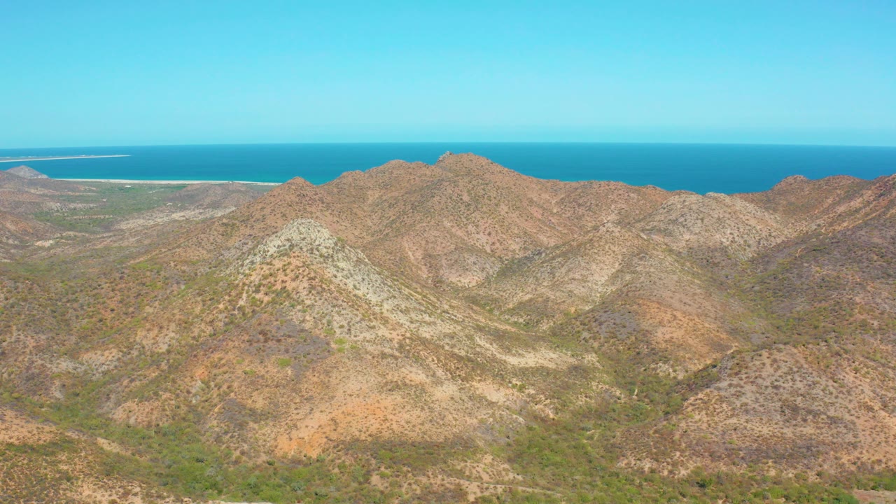 Aerial frame over Cabo Pulmo showing tan rocky mountain ridges, sparse green vegetation, a blue ocean horizon, and no visible people.