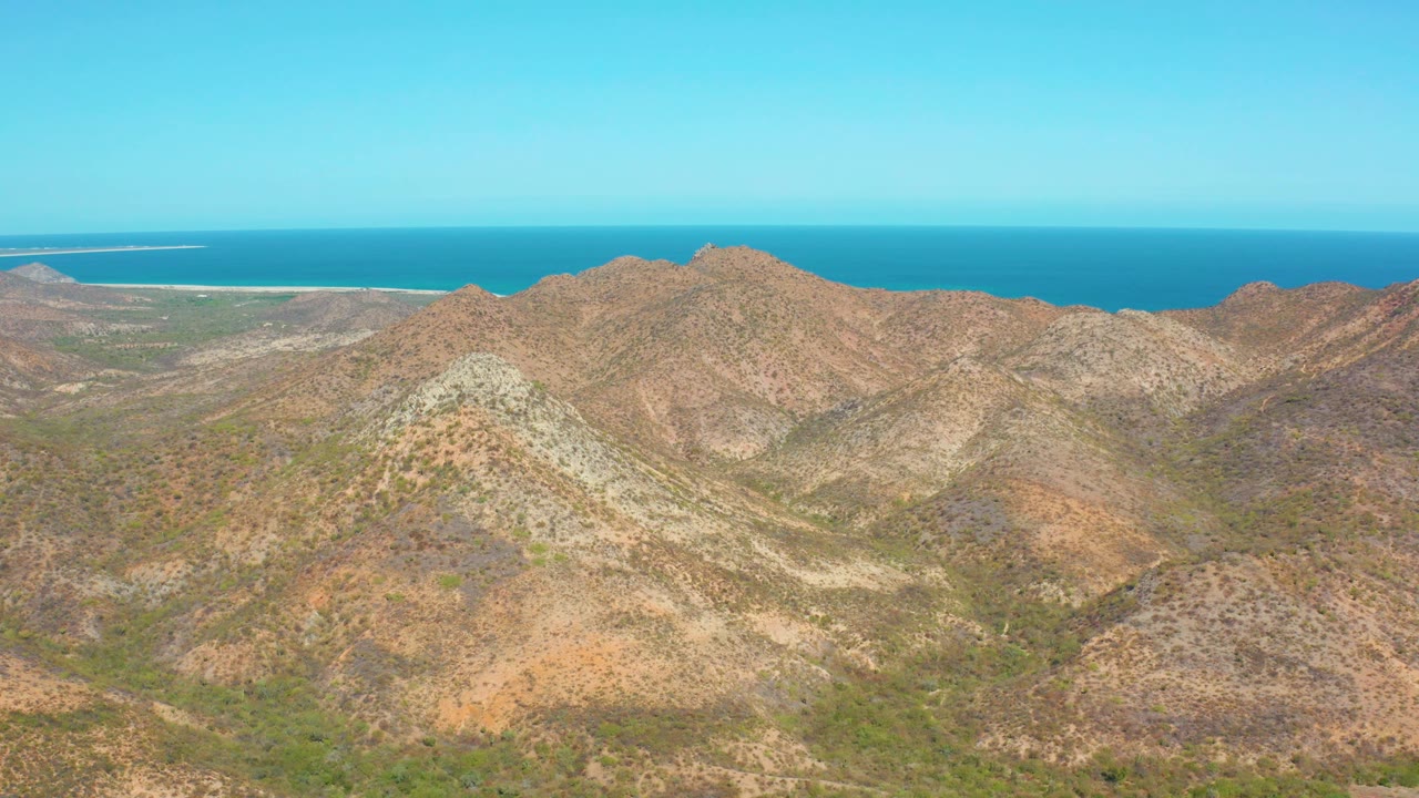 Aerial frame from Cabo Pulmo showing layered tan and brown mountain slopes, small patches of green vegetation, distant coastline, and no visible people.