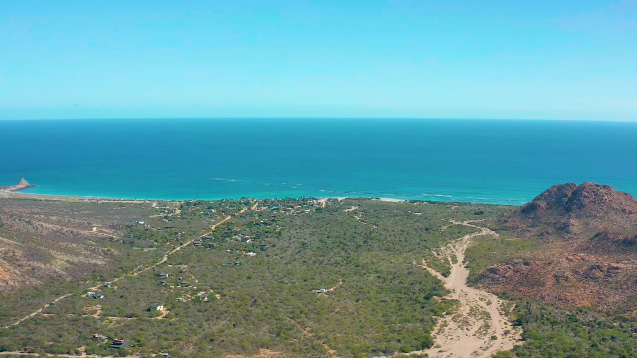 Opening aerial frame over Cabo Pulmo, Baja California Sur, showing green coastal dunes, pale dirt tracks, rocky hills to the right, turquoise ocean on the horizon, and no visible people.