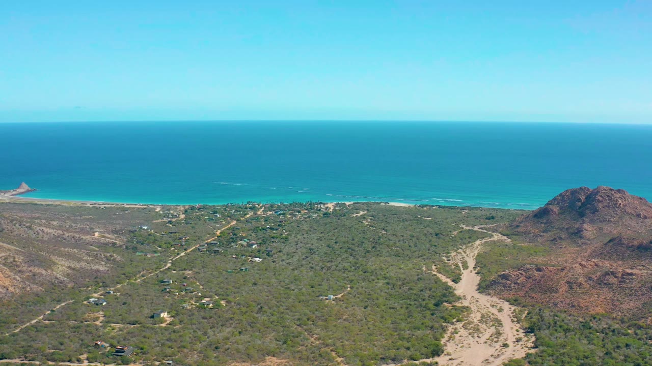 Wide drone view of coastal dunes and a rocky hill beside blue ocean.