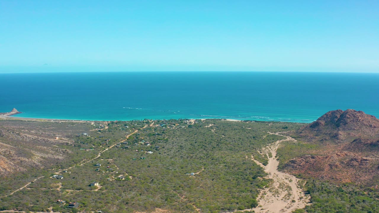 A high aerial view over Cabo Pulmo shows intersecting dirt tracks, mixed green and dry terrain, the reddish hill mass to the right, the ocean beyond, and no visible people.