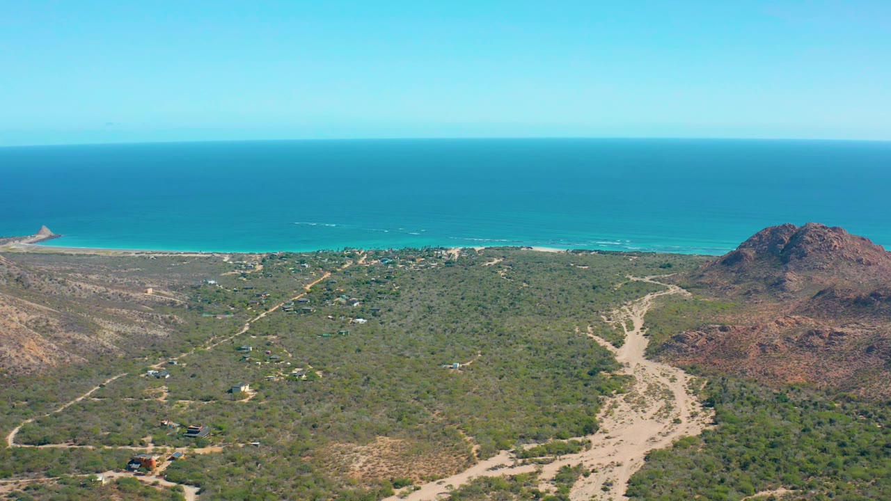 Wide drone view of coastal vegetation, dirt tracks, rocky hill, and ocean horizon.