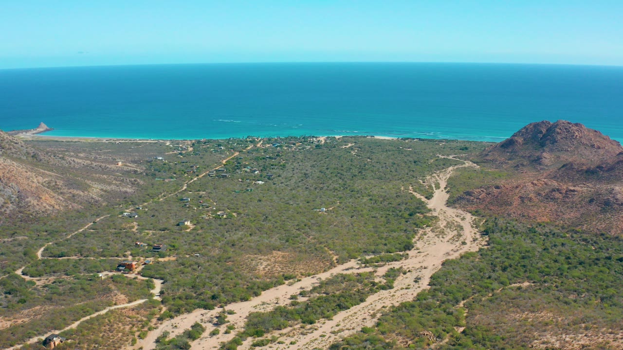 At 0:00, an aerial view over Cabo Pulmo shows scrub-covered hills, pale sandy washes, a reddish rocky hill at right, turquoise ocean beyond the beach, and no visible people.