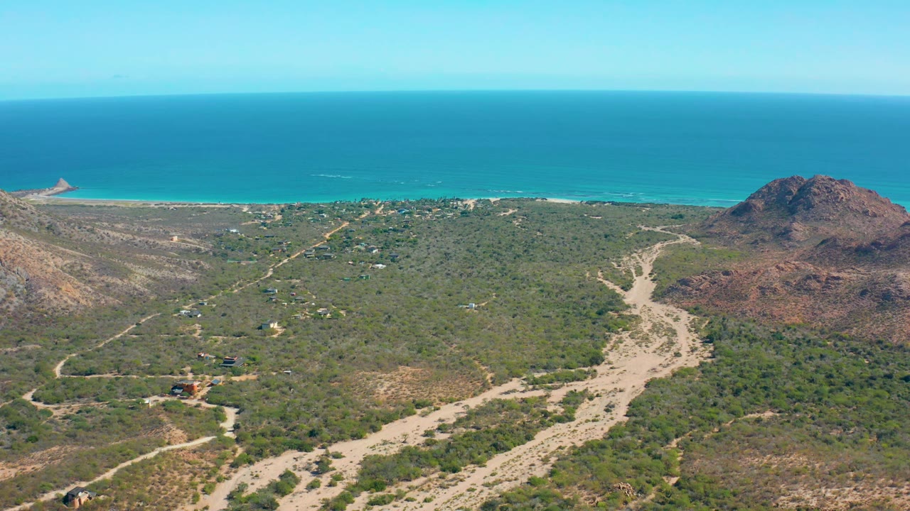 At 0:01, the Cabo Pulmo coastline remains wide in frame, with sandy tracks crossing green scrub, the rocky hill shifted near the right side, bright ocean behind it, and no visible people.