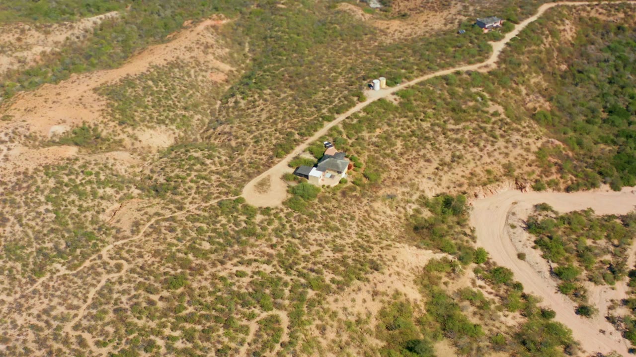 High aerial frame over Cabo Pulmo, Baja California Sur, showing a winding sandy road, patchy green scrub, dry hills, and small buildings with no clearly visible person at this moment.