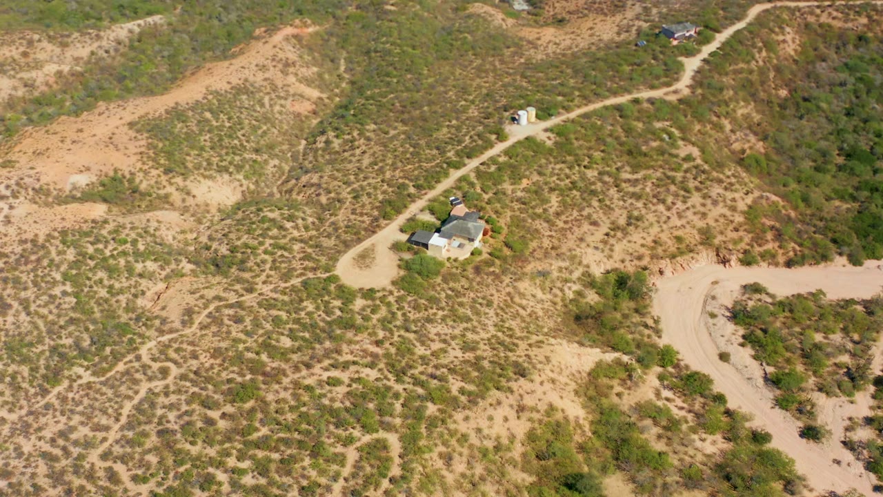 Drone view of a winding dirt track, scrubby hillside, and a small structure.