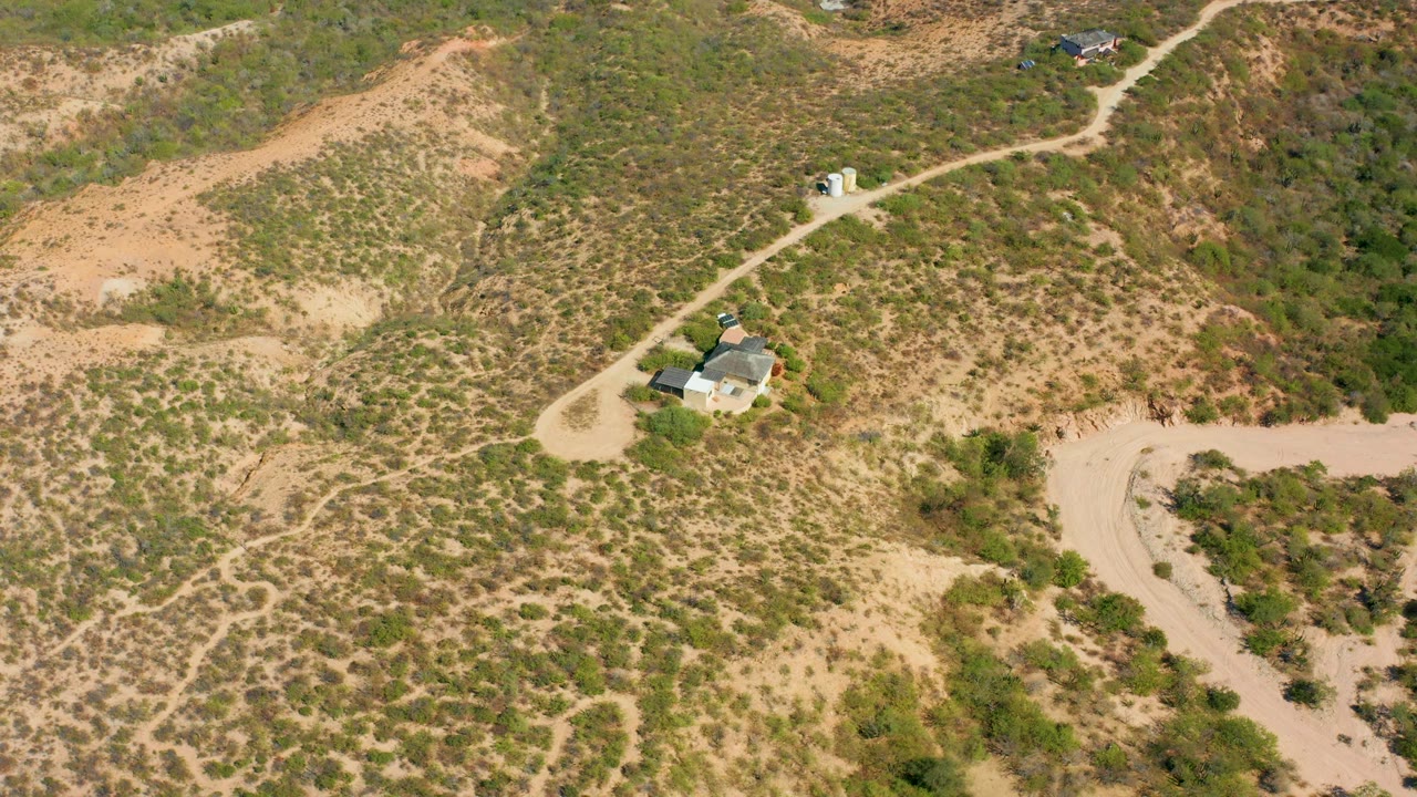 High drone frame over Cabo Pulmo dry terrain, with a sandy road running through low scrub, a small building cluster, and open hillside filling the frame.