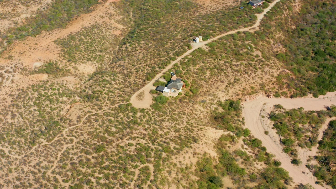 Aerial view in Cabo Pulmo, Baja California Sur, showing a winding dirt road across dry scrub-covered hills, a small vehicle near the upper road, a light-colored house below it, and no visible people.