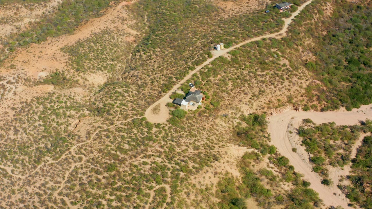 Drone view over Cabo Pulmo, Baja California Sur, where the dirt road curves through scrubby hills, a small vehicle sits near the upper road segment, a house is visible near the road, and no visible people appear.