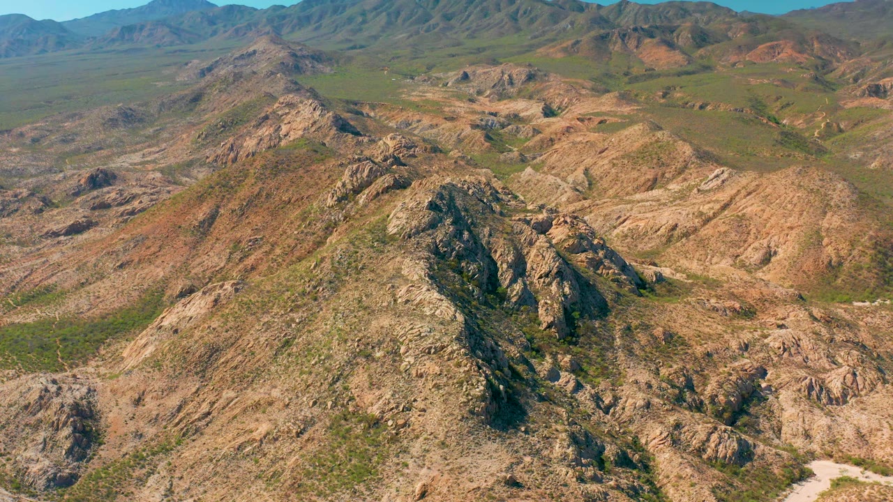 Aerial frame over Cabo Pulmo showing a central rocky ridge, tan eroded slopes, green scrub in the low areas, distant ridgelines, and no visible people.
