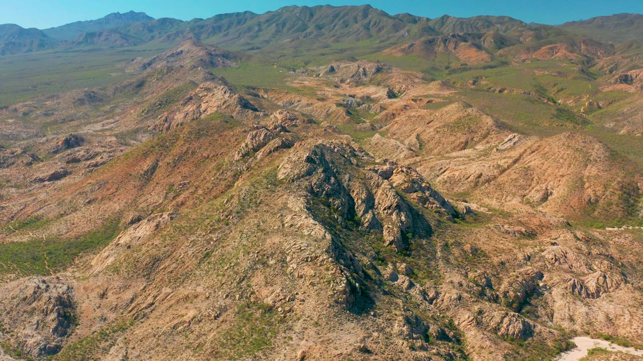 Mid-clip aerial frame over Cabo Pulmo arid hills, with a jagged rocky foreground, pale dry wash at the lower edge, green valley patches, distant mountains, and no visible people.