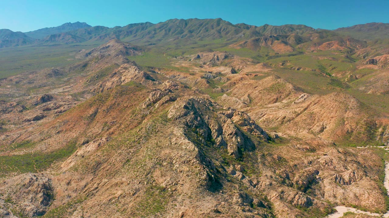High aerial frame over Cabo Pulmo with tan ridges in the foreground, green vegetation in the valley, layered mountains on the horizon, and no visible people.