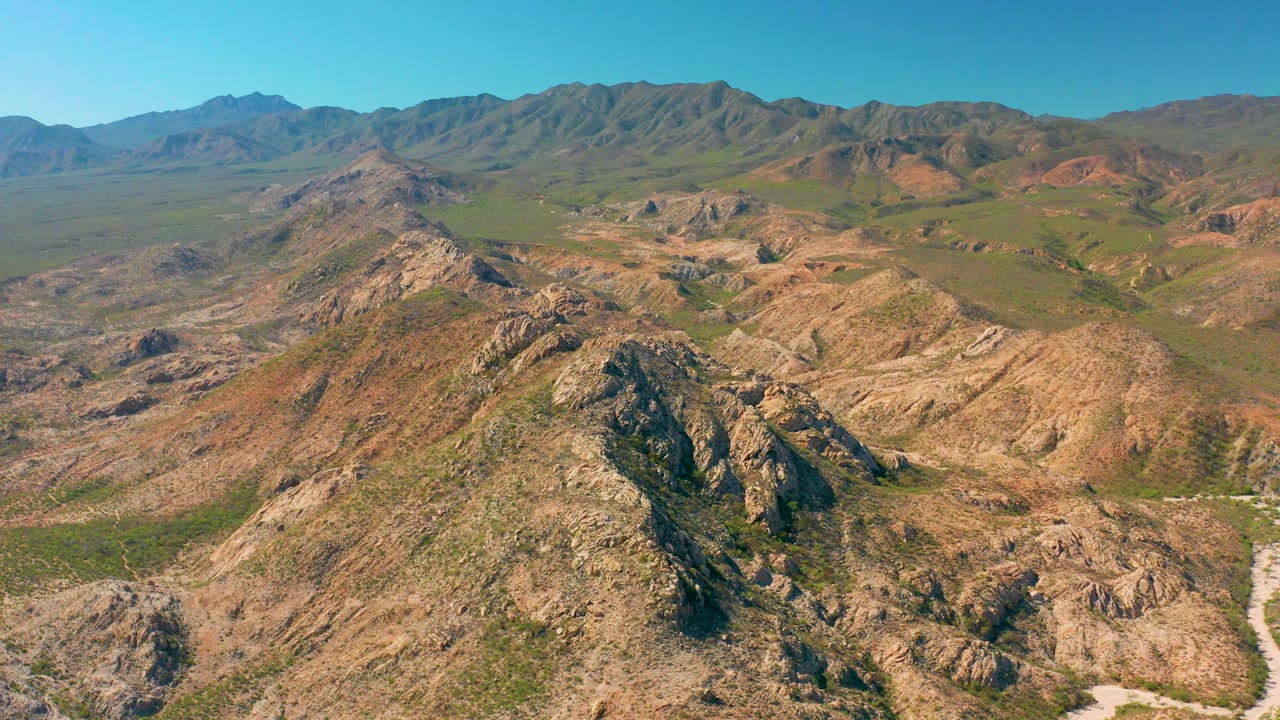 Cabo Pulmo aerial frame with rugged foreground ridges, green vegetation spread across the valley, a broad mountain backdrop, and no visible people.