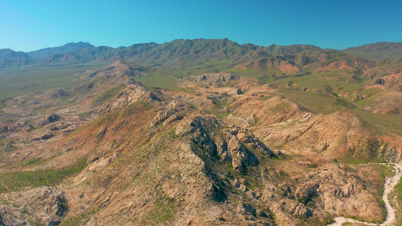 Aerial frame from Cabo Pulmo, Baja California Sur showing tan rocky ridges in the foreground, green desert valley beyond, a pale dirt road at right, distant mountains, and no visible people.