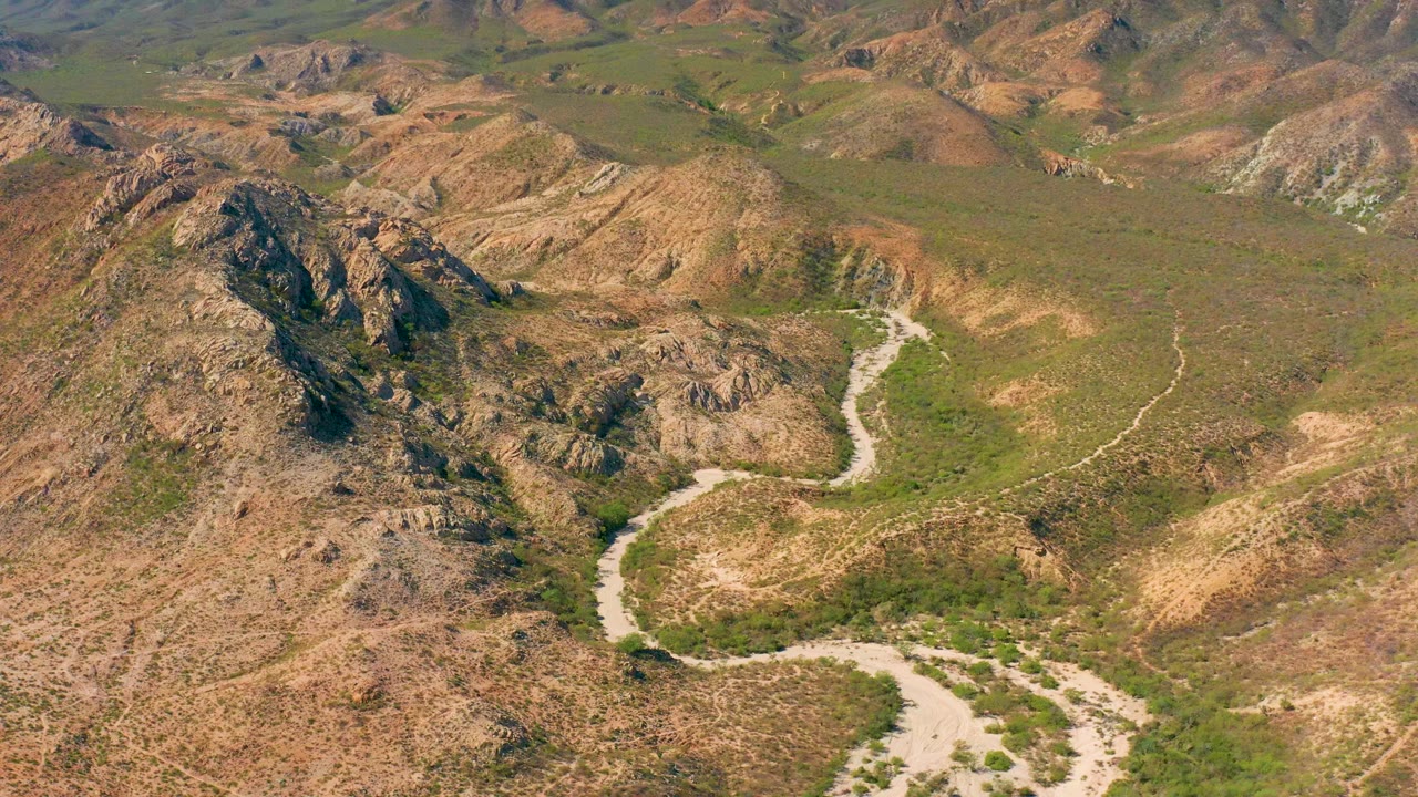Aerial frame over Cabo Pulmo, Baja California Sur showing a pale dirt road curving through rocky green-brown hills, with no visible people.