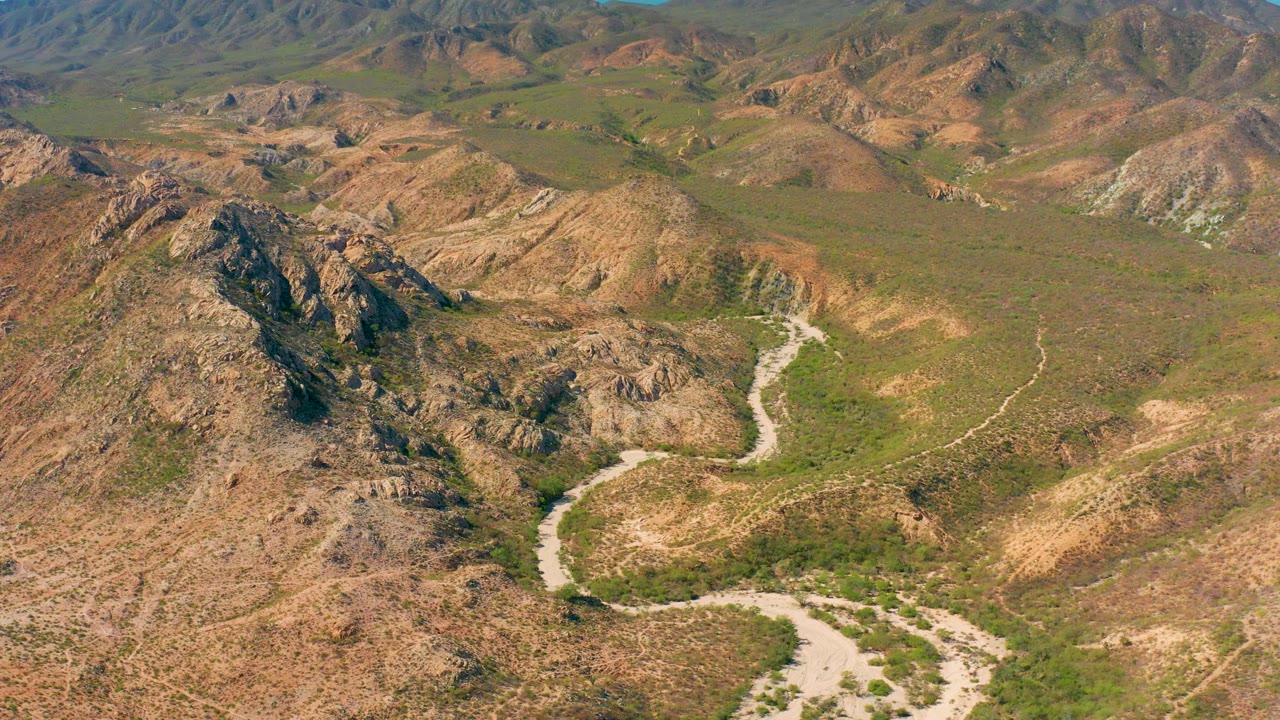 Aerial view in Cabo Pulmo with a pale river channel leading past dark rocky outcrops, scattered green scrub, layered brown hills, and no visible people.