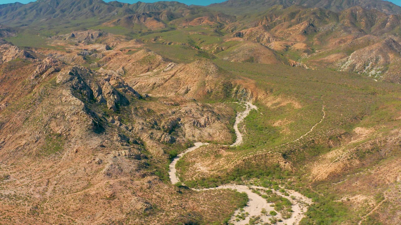 Cabo Pulmo aerial frame showing the pale wash bending through the lower valley, with jagged rocky hills in the foreground, green scrub in the basin, and no visible people.