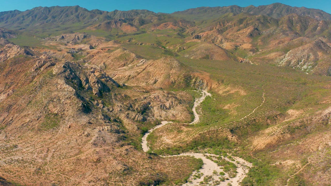 A high aerial frame over Cabo Pulmo shows a pale dirt road curving through rocky tan hills with green vegetation and no visible people.