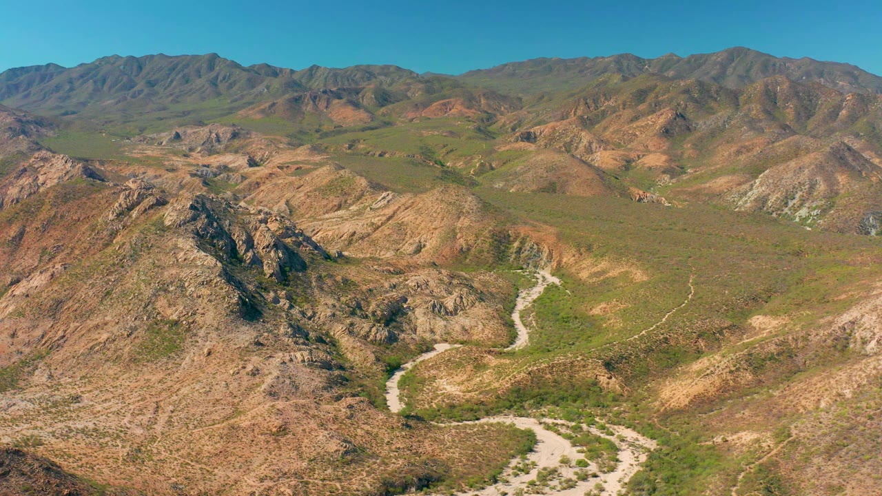 Early frame of the Cabo Pulmo valley showing the winding road lower in the frame, rugged hills on both sides, distant mountains, and no visible people.