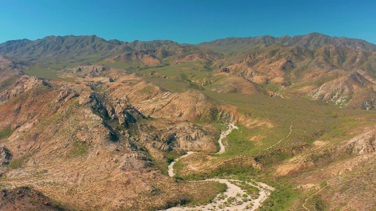 Aerial frame over Cabo Pulmo terrain with the winding pale track lower in frame, rocky ridges on both sides, green valley vegetation, distant mountains, and no visible people.