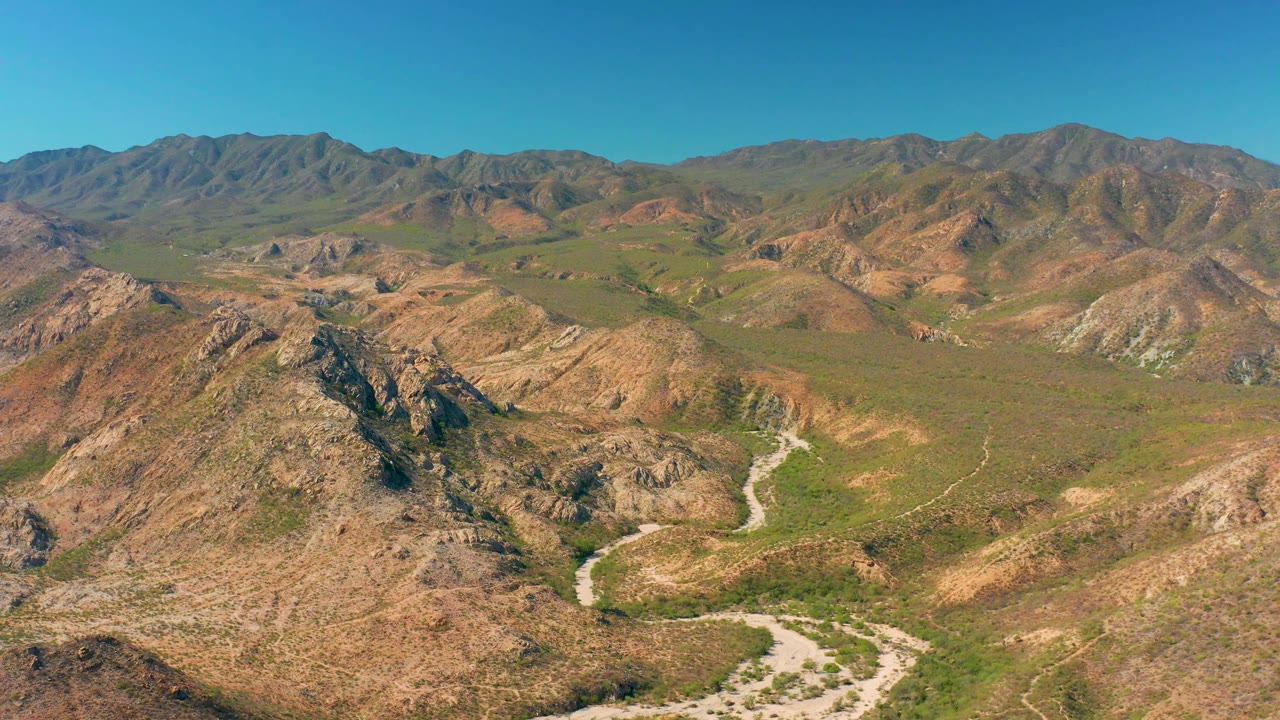Wide aerial frame from Cabo Pulmo showing sunlit brown slopes, patches of green vegetation, a pale route curving through the lower valley, distant mountains, and no visible people.