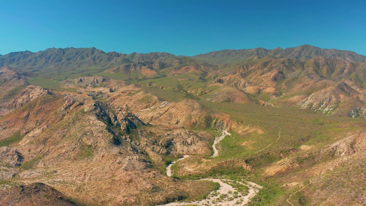 Wide aerial frame from Cabo Pulmo with the road more centered between tan rocky slopes, green low vegetation across the valley, distant ridgelines, and no visible people.