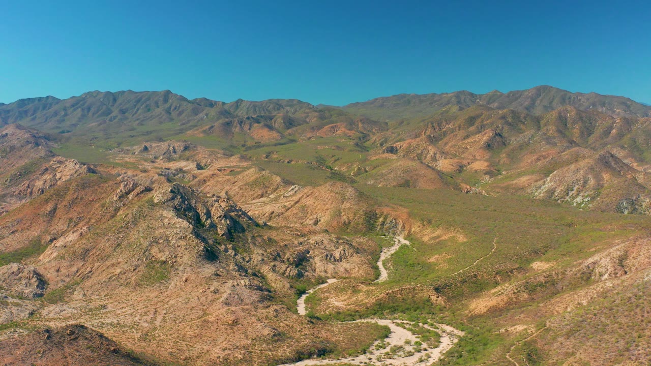 Drone vantage over Cabo Pulmo terrain with a pale road bending through the midground, rocky hills on both sides, a broad green valley floor, distant ridges, and no visible people.
