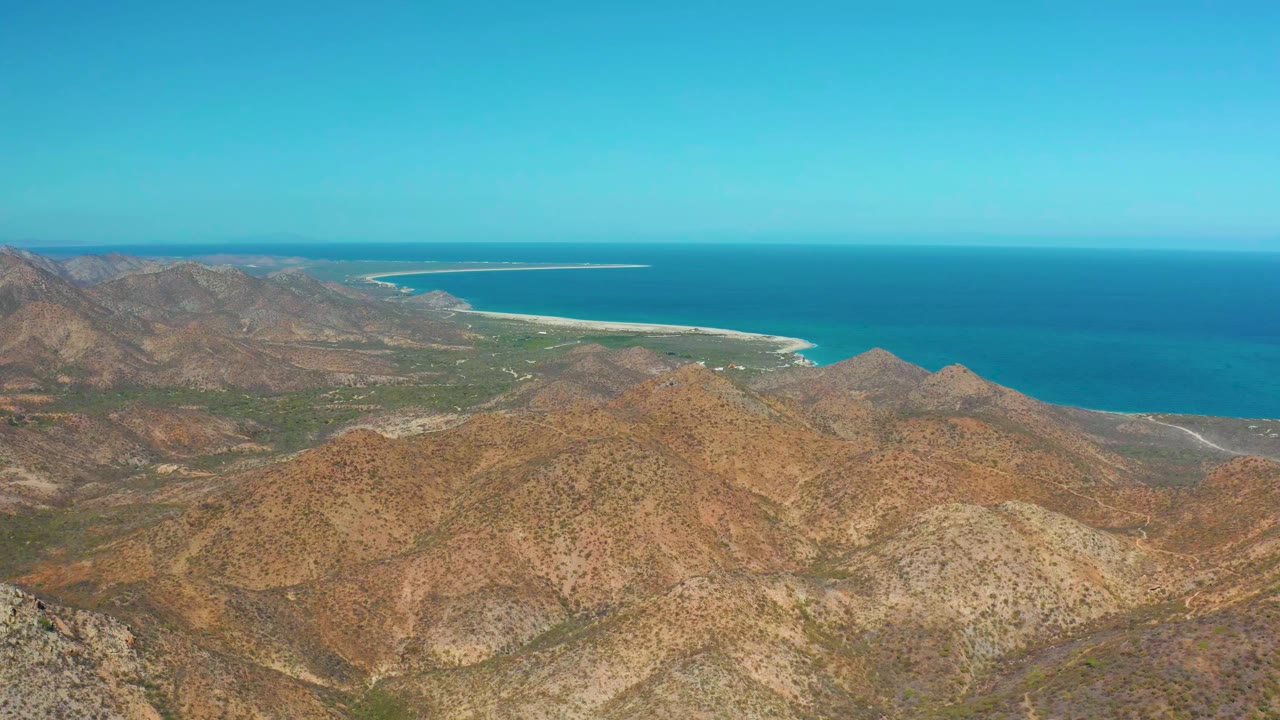 Cabo Pulmo aerial frame with tan mountain ridges across the lower view, a green coastal strip, a curved sandy beach, deep blue water, and no visible people.