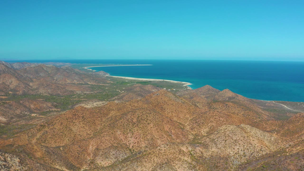 Mid-clip frame over Cabo Pulmo shows layered arid hills, a thin coastal road or track, pale surf line, deep blue ocean, clear sky, and no visible people.