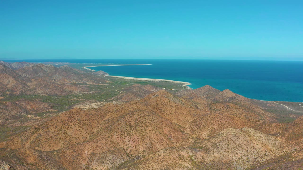 Cabo Pulmo aerial frame with brown hills in the foreground, a green strip near the shore, pale sand along the water, blue ocean, clear sky, and no visible people.