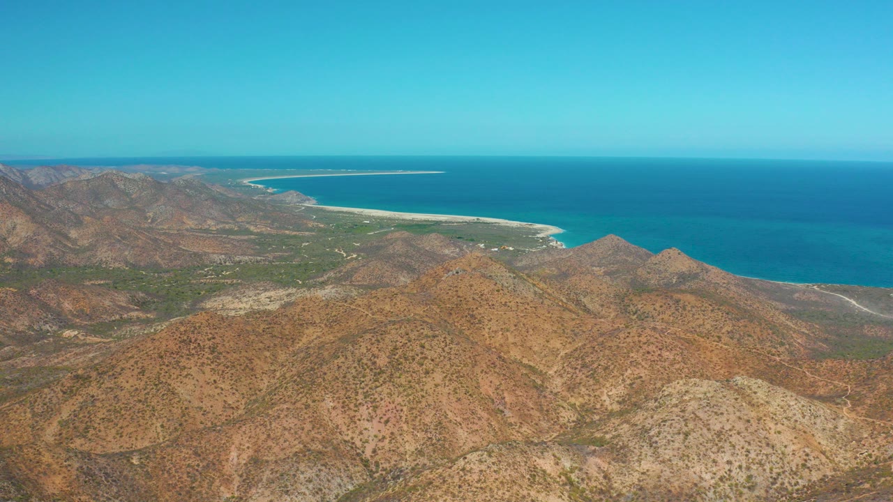Opening aerial frame over Cabo Pulmo, Baja California Sur with dry hills in the foreground, a pale beach and blue ocean beyond, and no visible people.
