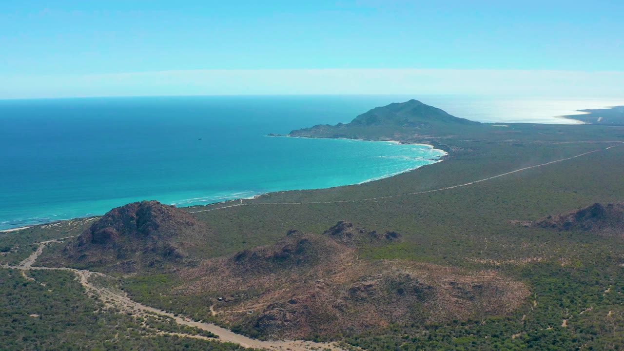 Wide aerial view of Cabo Pulmo with turquoise ocean on the left, a sandy curved shore, dry foreground hills, a pale road through scrubland, a distant headland, and no visible people.