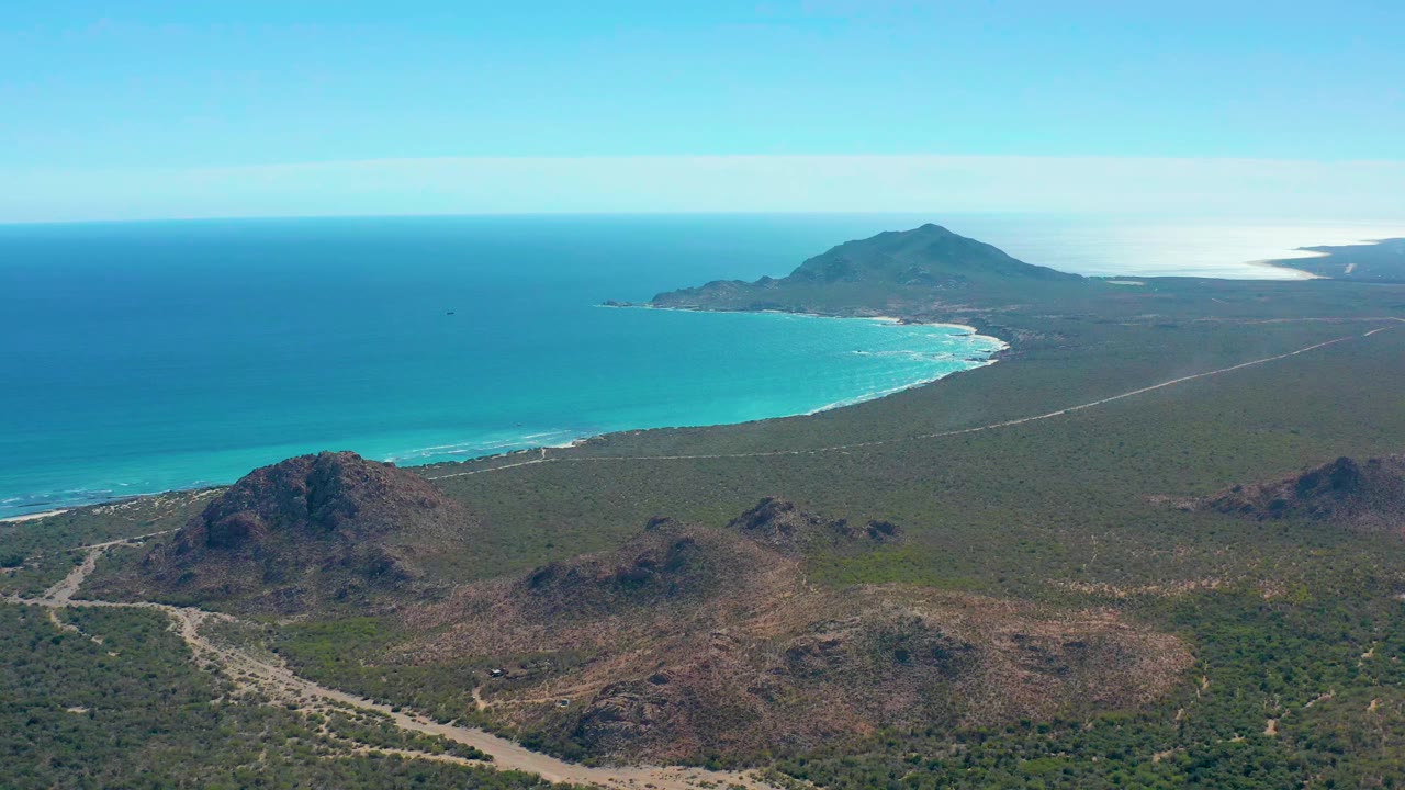 Aerial frame over Cabo Pulmo showing rocky brown hills in the foreground, a scrub-covered plain, the curved turquoise bay, a distant headland, bright sky, and no visible people.