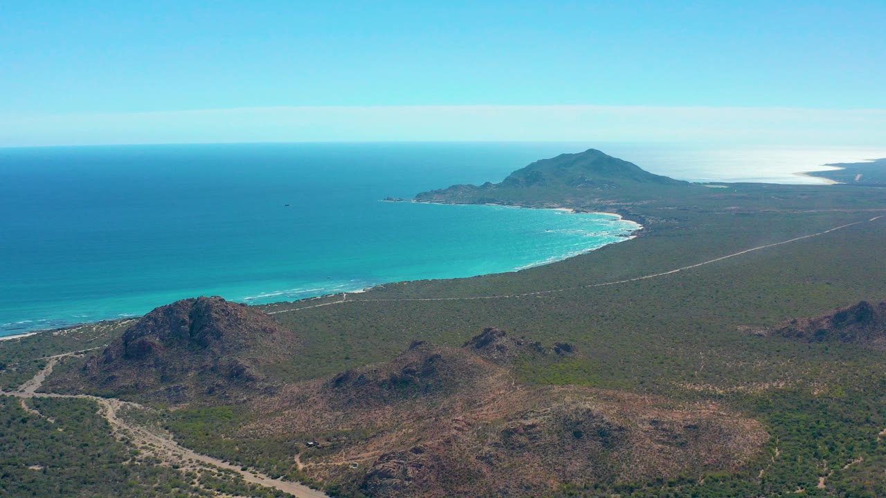 Wide aerial frame over Cabo Pulmo showing rocky brown hills in the foreground, sparse desert vegetation, a curved turquoise beach, deep blue ocean, distant mountains, and no visible people.
