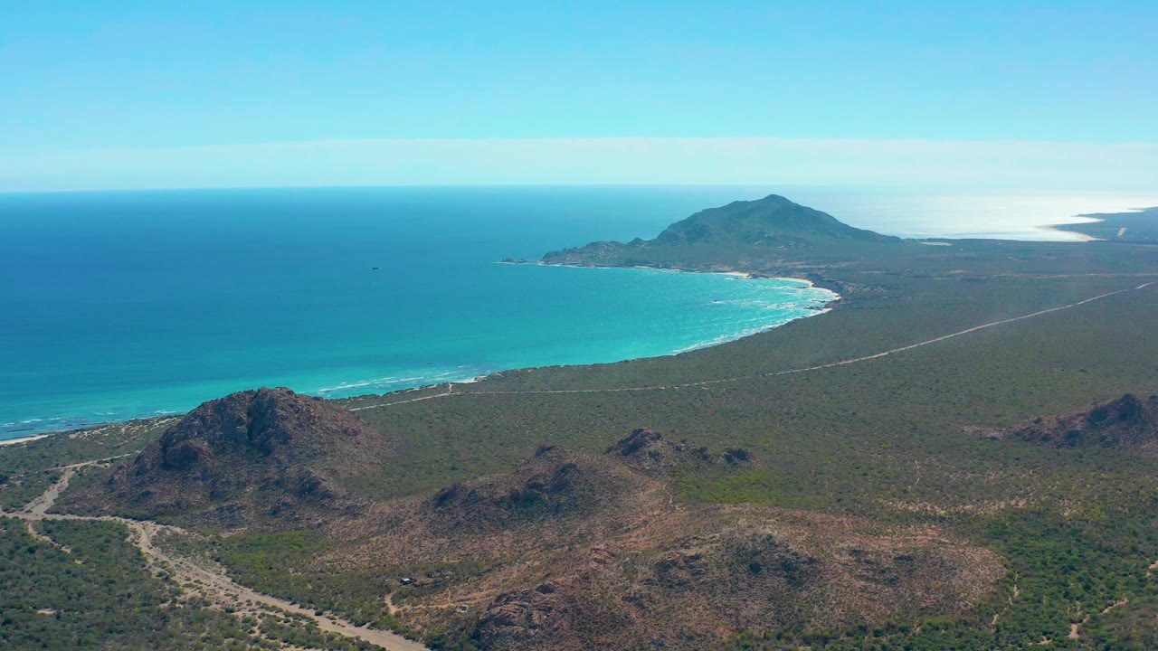 Cabo Pulmo aerial frame with a pale road crossing the scrubland, low rocky hills below, bright turquoise surf along the shore, a blue ocean horizon, and no visible people.