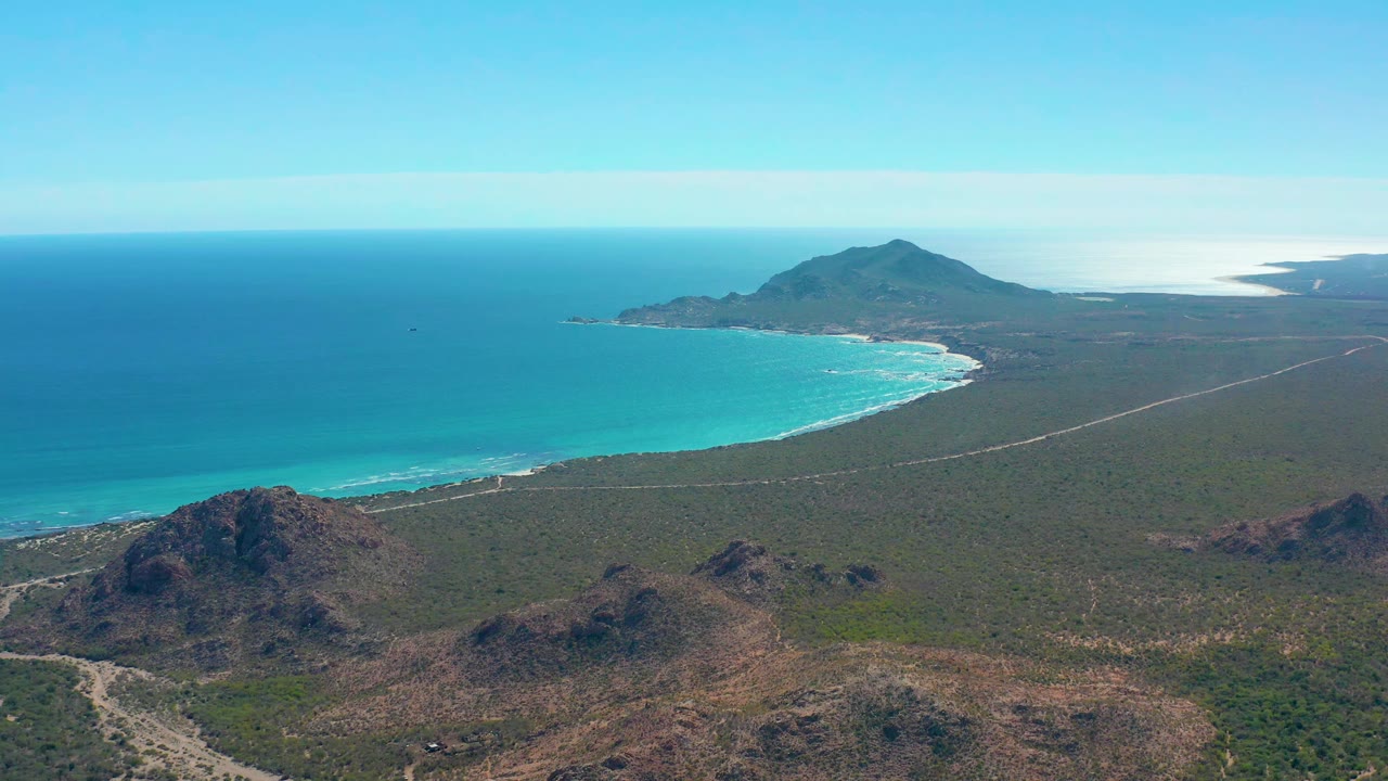 Aerial view in Cabo Pulmo with the turquoise shoreline and surf line in the left midground, rocky brown hills in front, a straight road across green scrub, and no visible people.