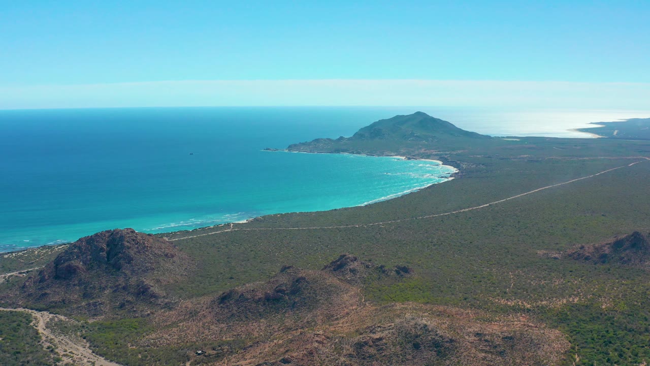 Cabo Pulmo aerial frame showing the bay stretching left to right, white surf along the beach, dry hills in the foreground, a distant headland under clear sky, and no visible people.