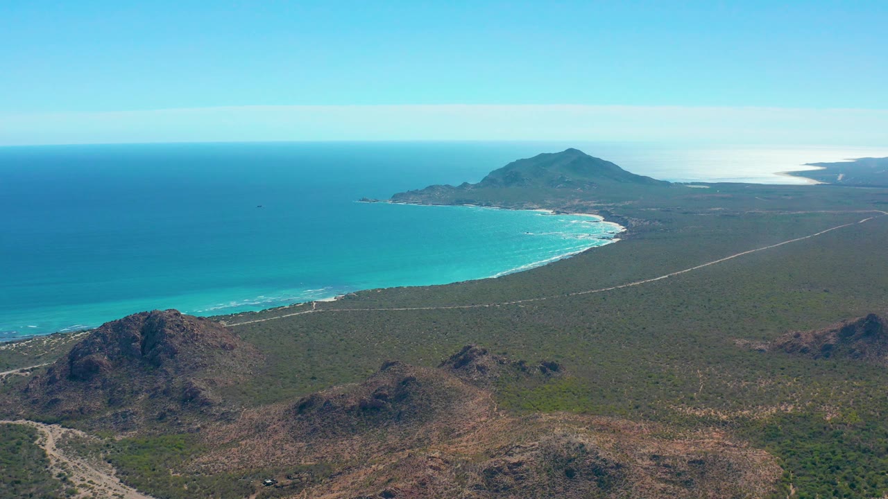 Aerial frame over Cabo Pulmo with calm blue ocean on the left, a pale sandy beach below the surf, scrub-covered flatland crossed by a road, foreground rock hills, and no visible people.