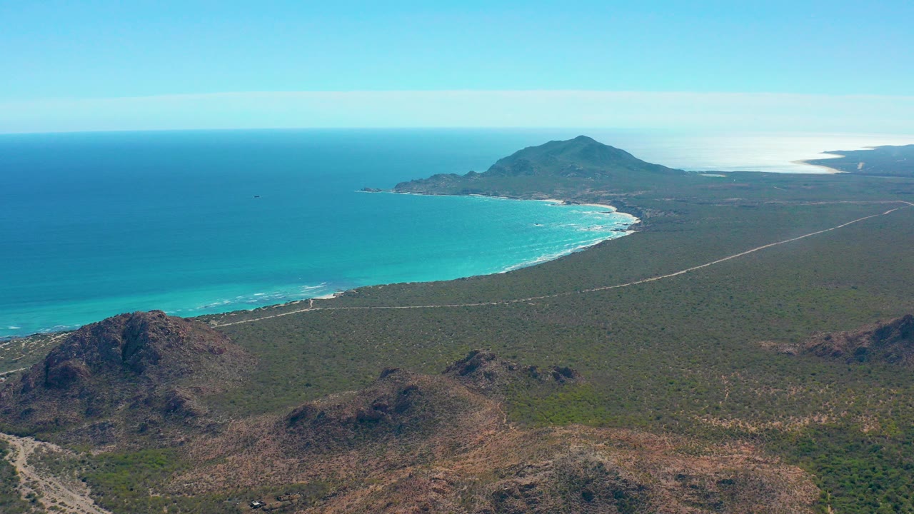 Opening aerial frame over Cabo Pulmo, Mexico, showing turquoise ocean, pale surf, sandy beach, a dirt road through semi-arid terrain, rocky hills in front, and no visible people.