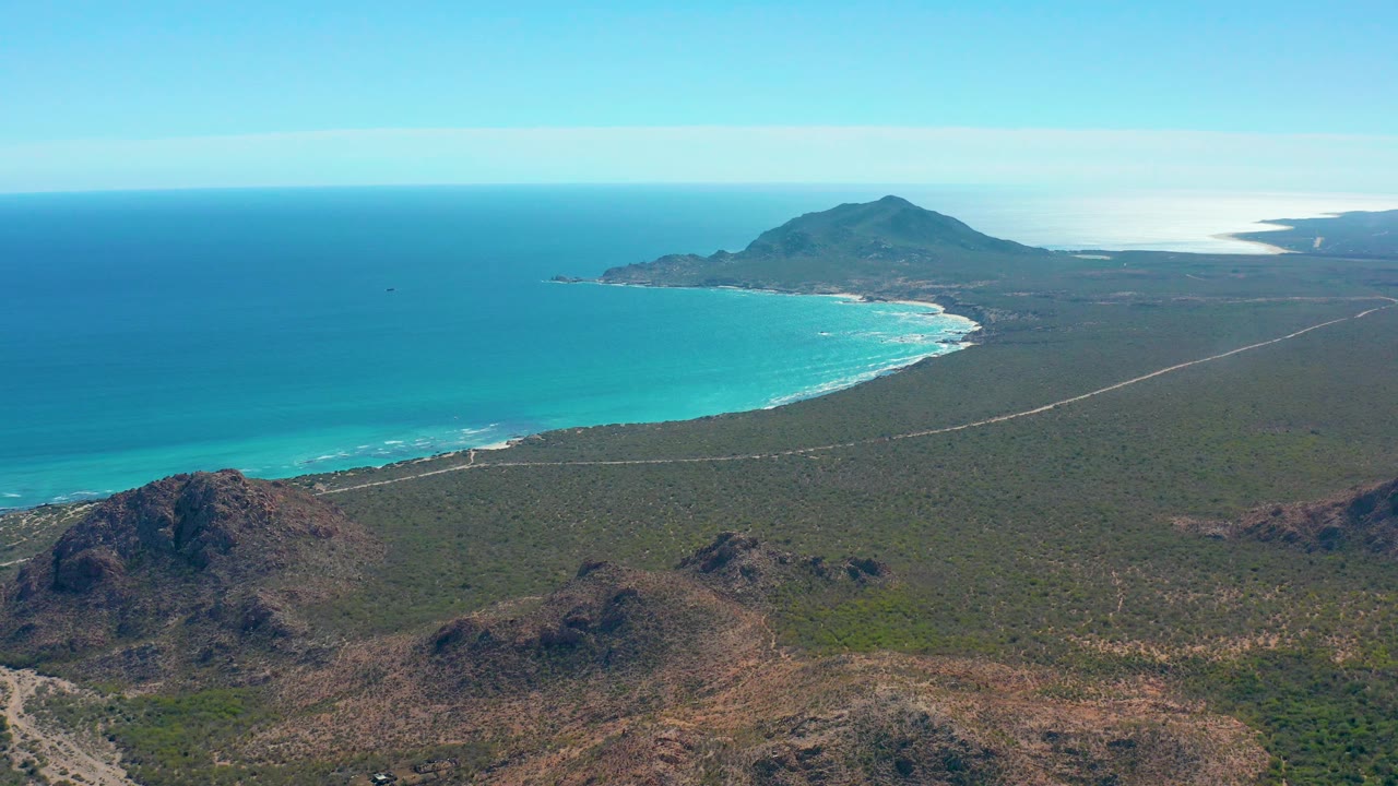 Coastal road beside blue ocean and low rocky desert hills.