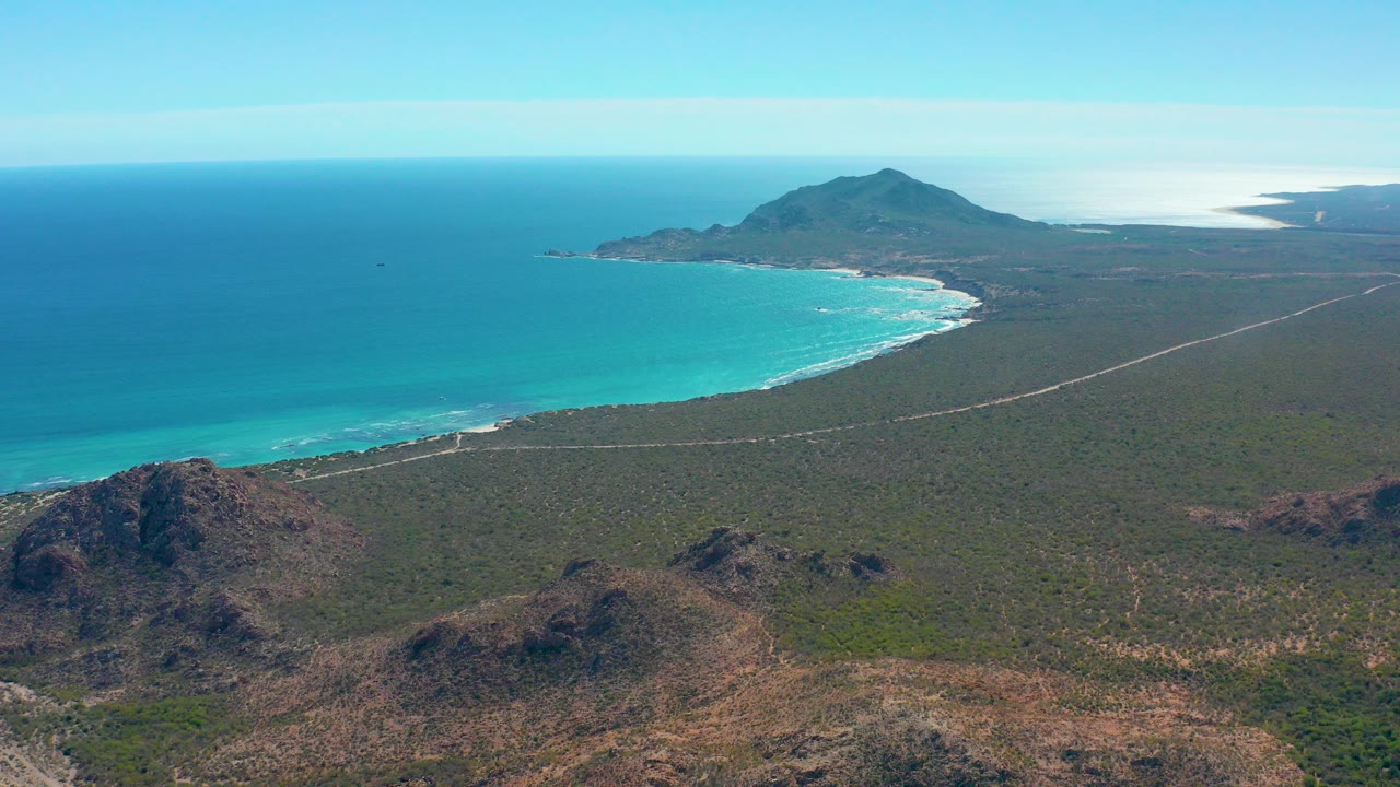 A wide aerial view over Cabo Pulmo shows turquoise water, a curved sandy beach, a pale road across green desert terrain, rocky foreground hills, and no visible people.