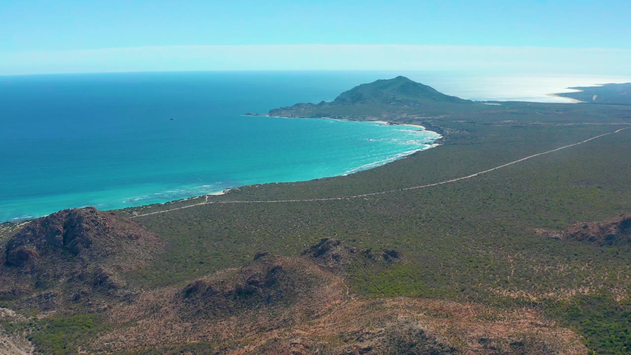 Aerial coastline with bright turquoise water, surf, desert scrub, and distant mountain.