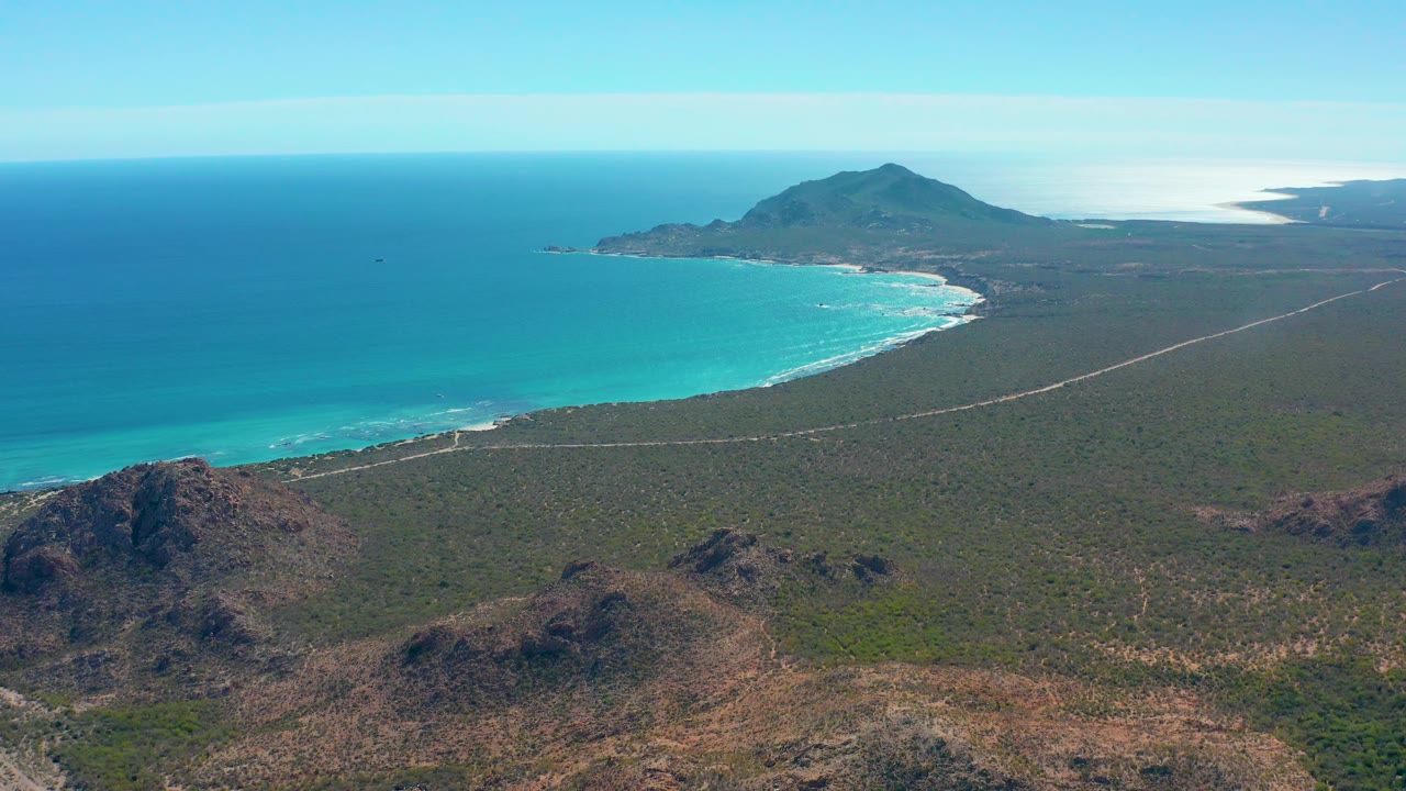 At 0:02, the aerial view shows Cabo Pulmo's turquoise bay, thin surf lines along the beach, scrub-covered terrain cut by a pale road, rocky ridges below, and no visible people.