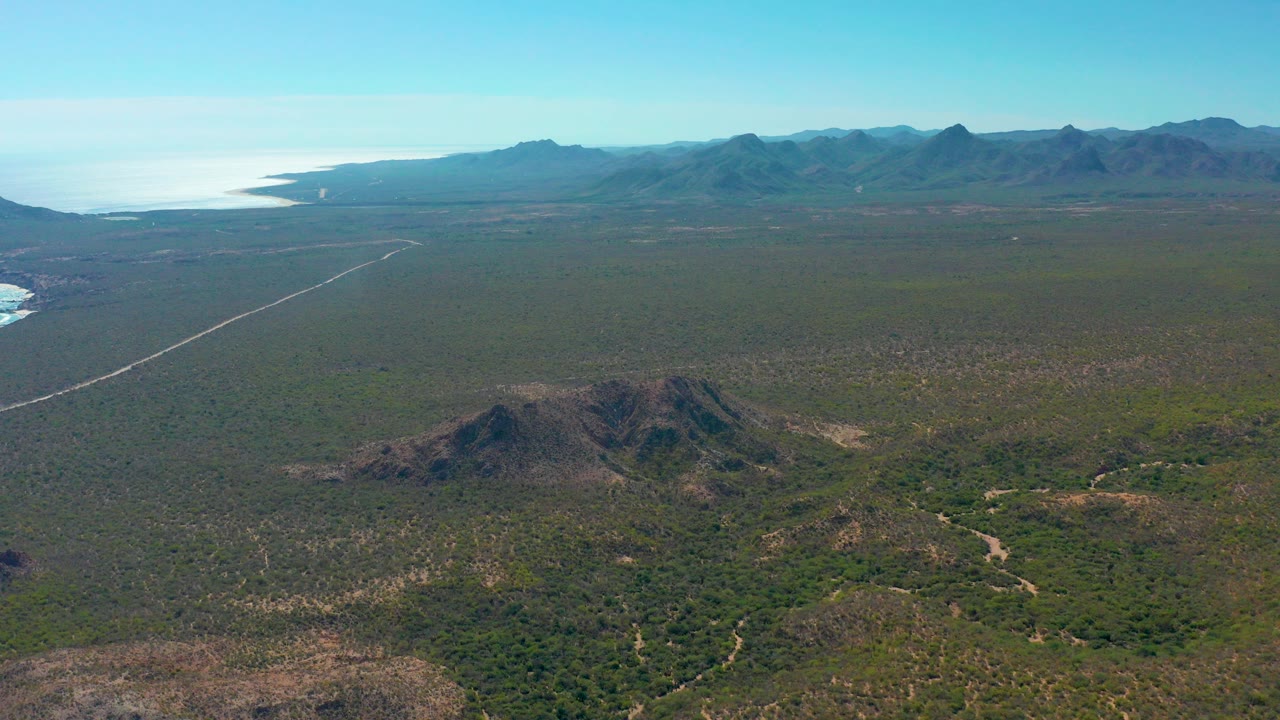 Aerial view in Cabo Pulmo, Baja California Sur, with a straight pale road crossing green-brown scrubland toward distant mountains and ocean at the left, no visible people.