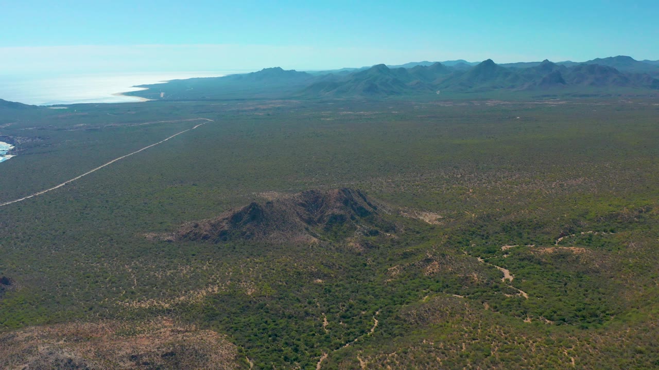 Wide aerial over Cabo Pulmo terrain with scrub-covered flats, a pale road running diagonally from the left, low hills below, mountains across the horizon, and coastline to the far left, no visible people.