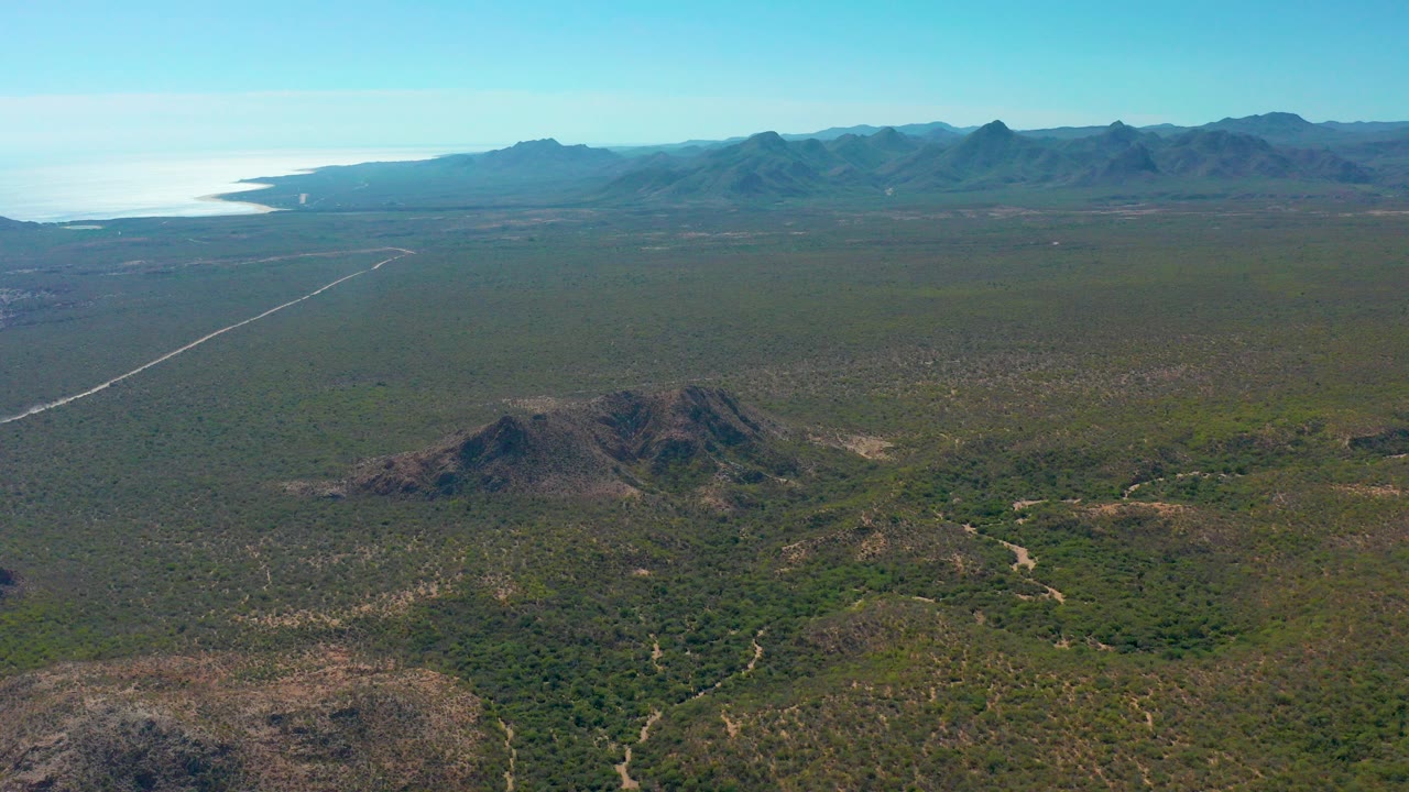 A high aerial view over Cabo Pulmo scrubland shows rocky foreground hills, a straight pale road at left, distant mountains, ocean on the horizon, and no visible people.