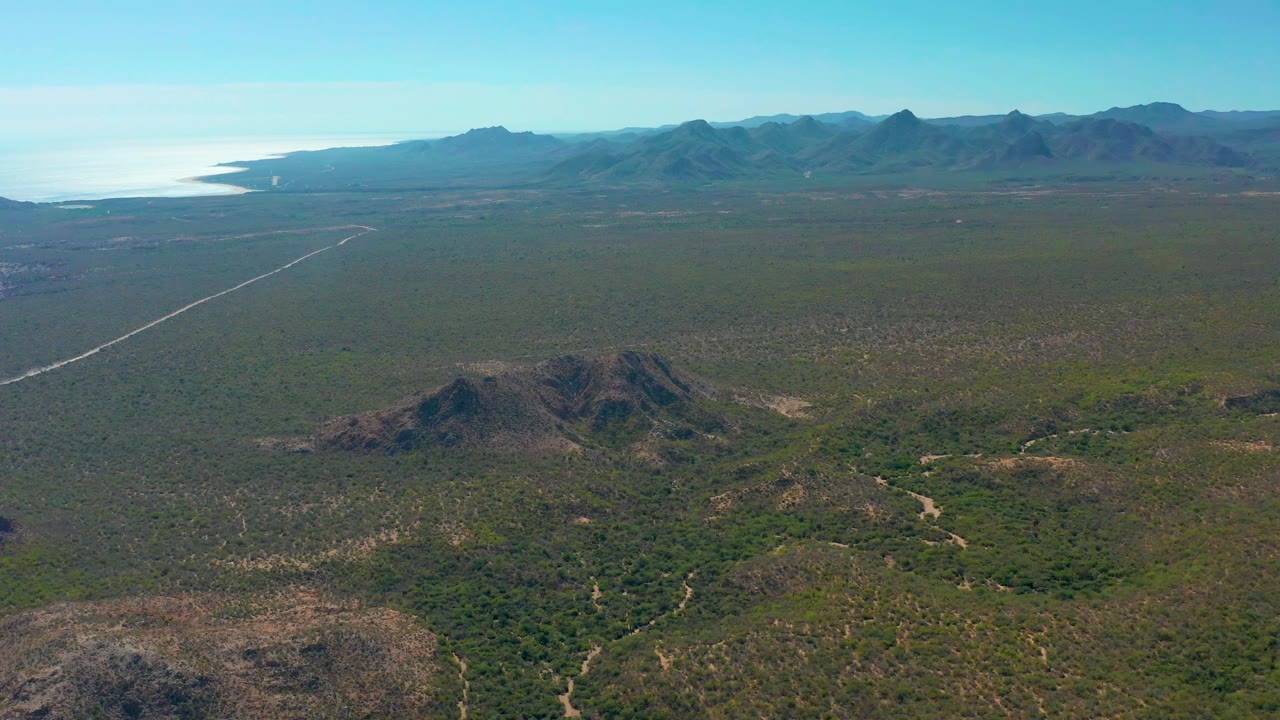 The Cabo Pulmo aerial remains above green scrubland, with low rocky hills centered below, the pale road running toward the left horizon, distant mountains, ocean, and no visible people.