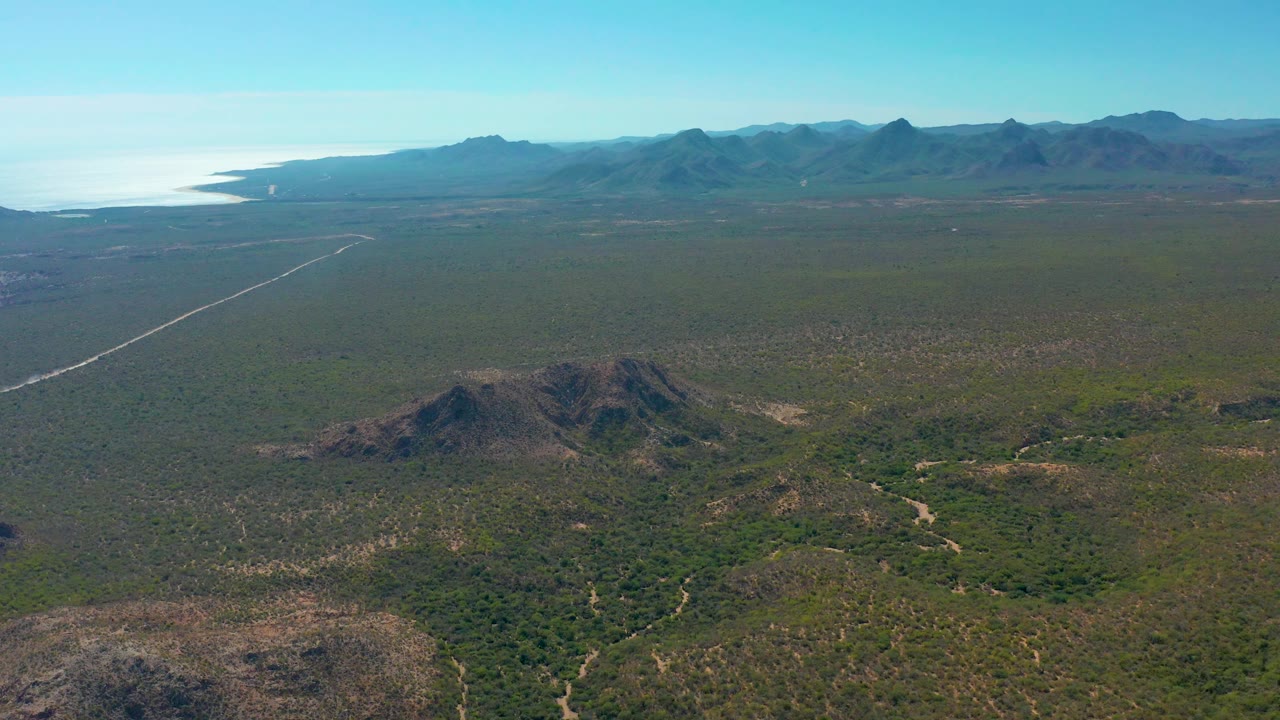 A wide Cabo Pulmo frame shows the coastal plain stretching to distant mountains, a straight road cutting through the left side, a bright ocean band beyond the land, and no visible people.
