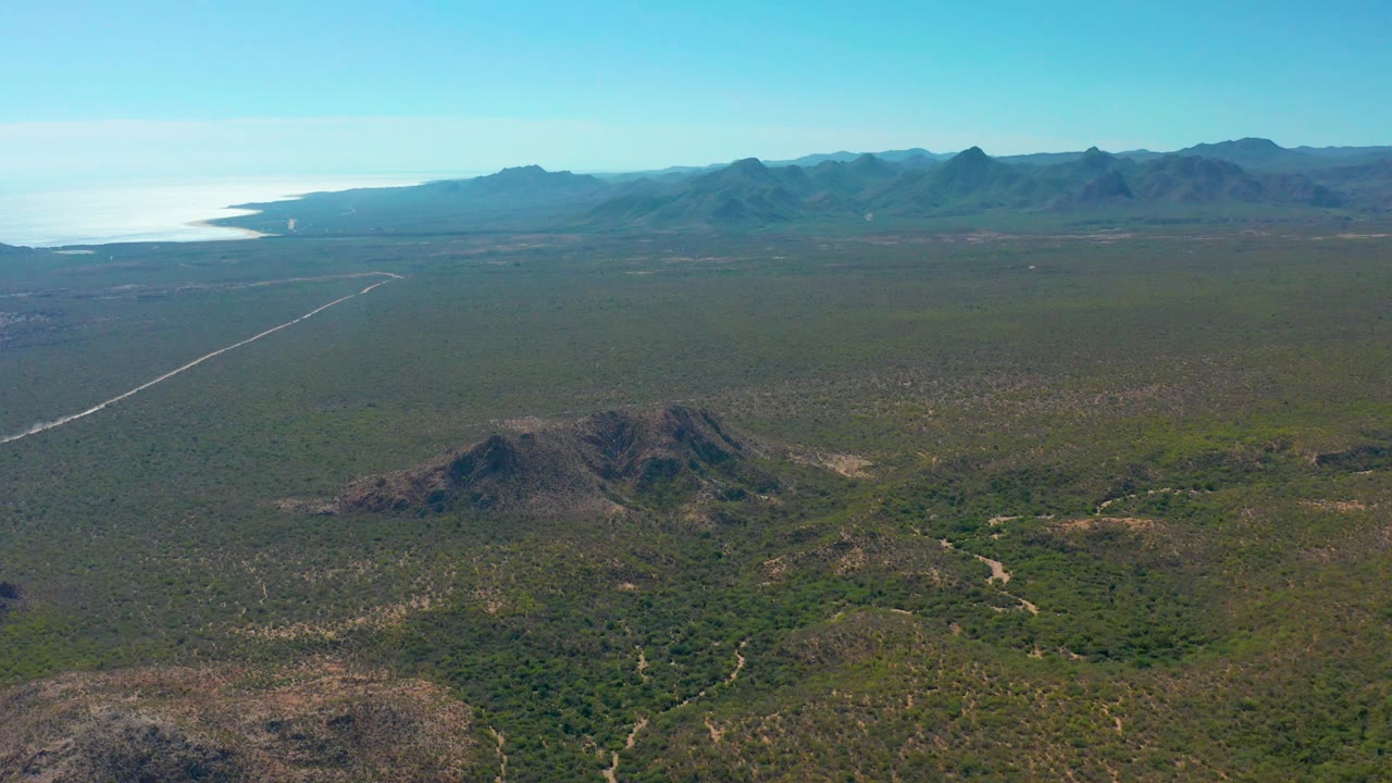 Aerial landscape with foreground hill, scrubland, road, ocean, and mountains.
